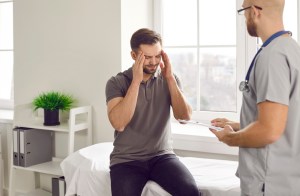 Male patient with terrible headache visiting his doctor. Young man suffering from migraine sitting on medical bed at clinic, holding his head and asking professional physician for help. Pain concept