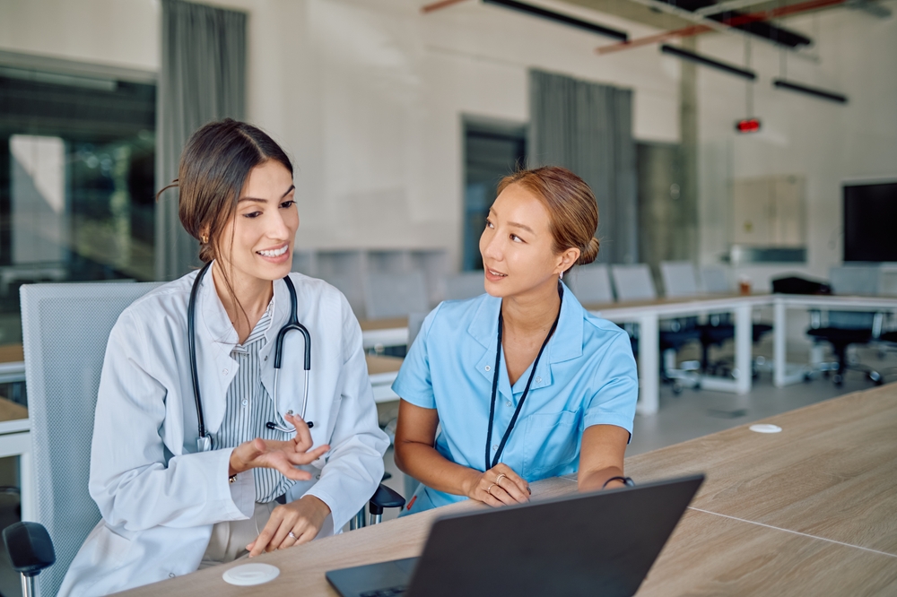 A female doctor and a female nurse are sitting at a desk in a hospital meeting room, discussing and using a laptop